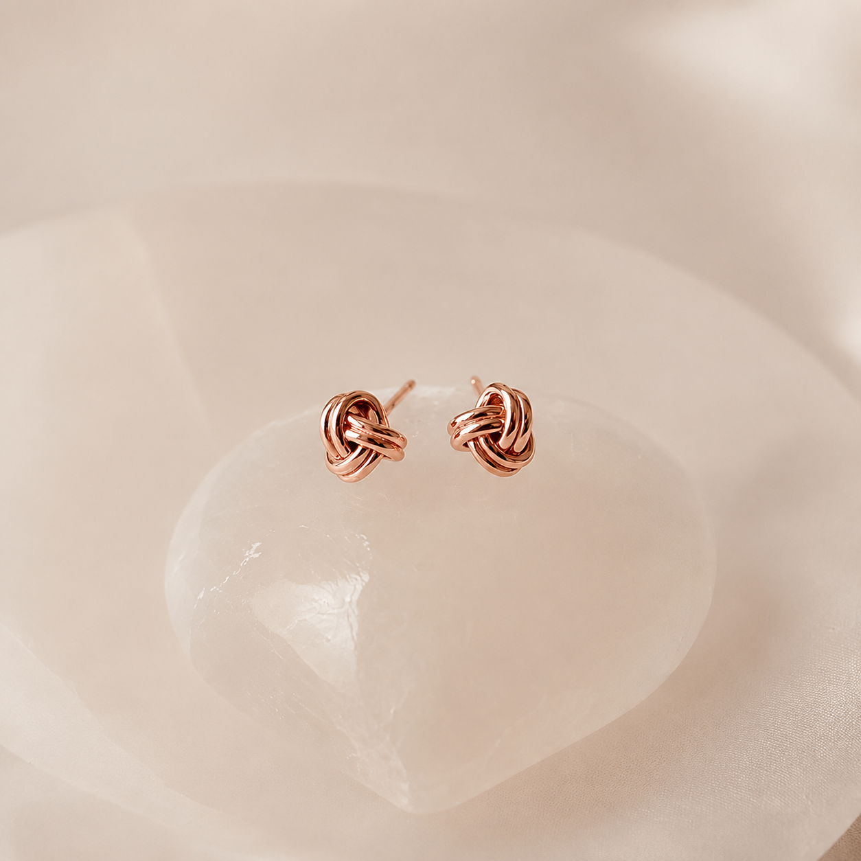 A pair of small rose gold knot stud earrings resting on a smooth, pale pink stone against a soft, warm beige background.